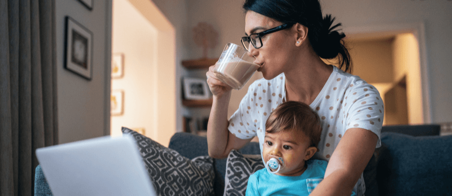 Mom drinking coffee and holding infant while looking at laptop